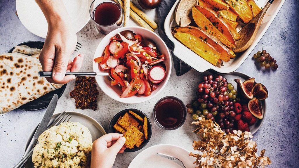 A vegan or vegetarian lunch table featuring plates of baked sweet potato, cauliflower, fruits, vegetable salad and tortilla with greens on a white background.