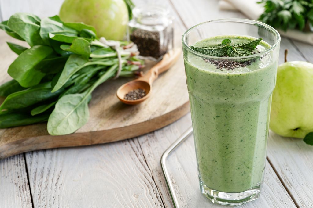 Green smoothie with fresh leaf vegetables, fruits, herbs, and chia seeds (spinach, green apple, parsley, mint, tarragon) in glass with reusable metal straw on white wooden table, healthy food