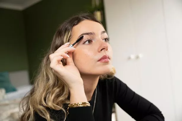 Three-quarter front view of woman with two-toned long hair wearing black top and gold jewelry, looking in off-camera mirror while putting on makeup.